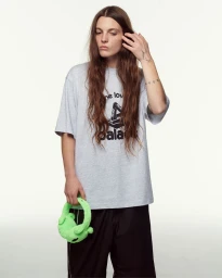 Model with long brown hair wearing a grey graphic t-shirt and black trousers holding bright green earmuffs.