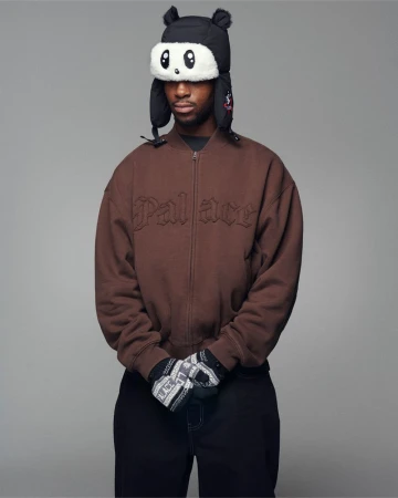 Man wearing a brown zip-up jacket, black beanie with panda ear flaps, and black trousers against a neutral grey studio backdrop.
