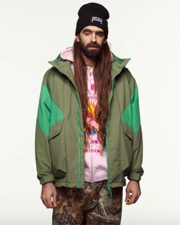 Man with long hair and beard wearing an olive green jacket, pink hoodie, black beanie, and camo trousers against a white background