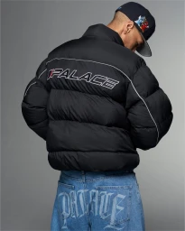 Man wearing a black puffer jacket with Palace branding and blue jeans, viewed from behind against a plain grey studio background.