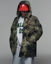 Model wearing a camouflage hooded jacket, red baseball cap, and logo sweatshirt against a plain grey studio backdrop.