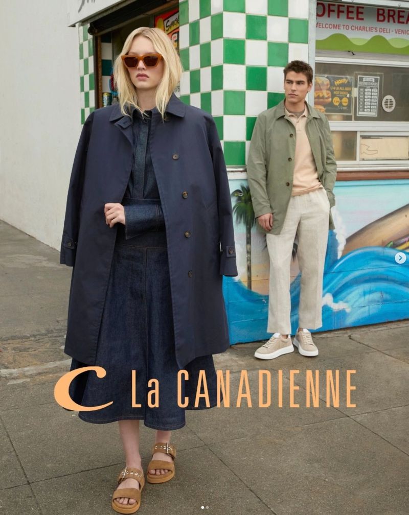 Two fashion models in an urban setting; a woman in a trench coat and denim dress stands next to a man in a light green jacket.