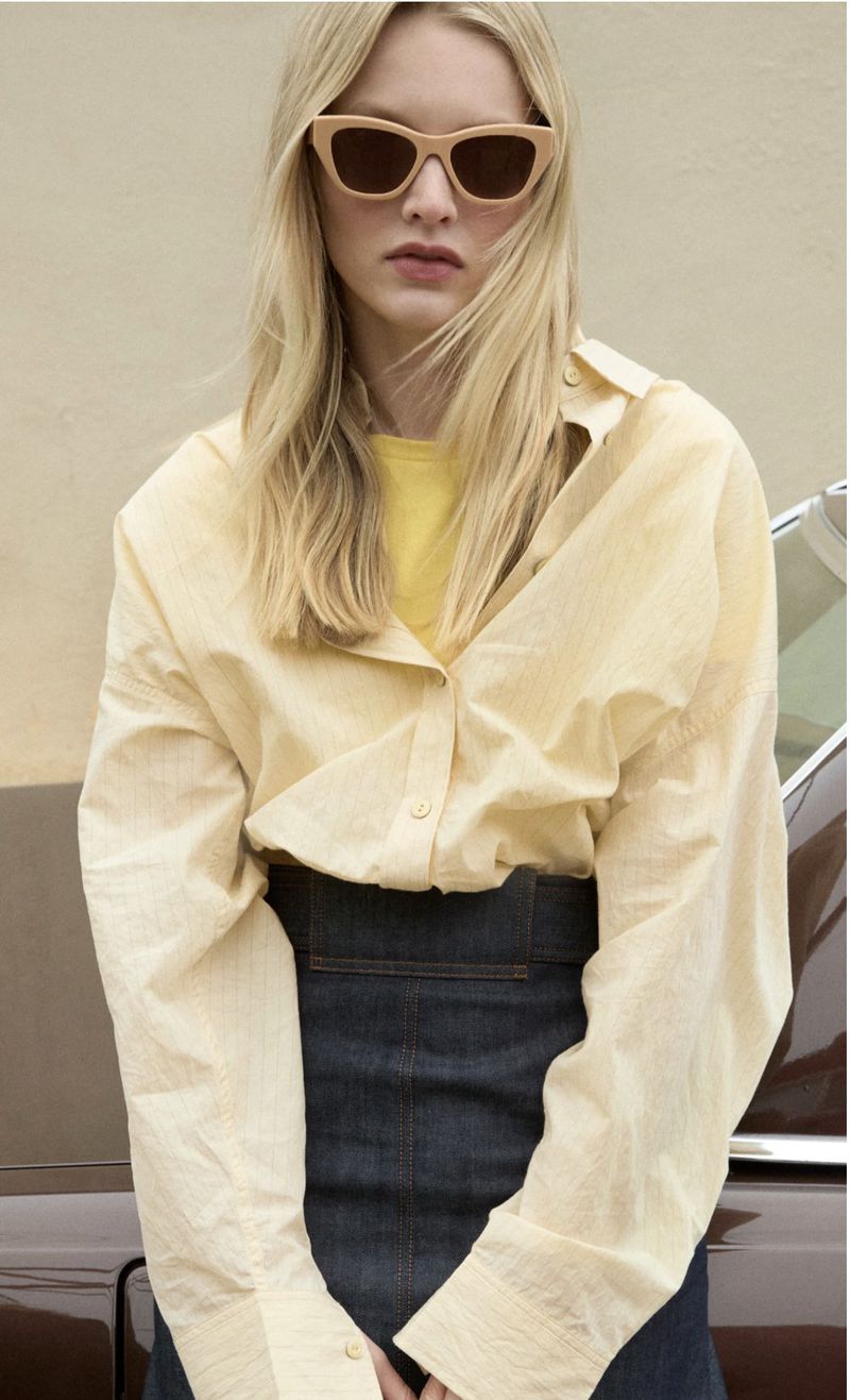 Model wearing oversized light yellow button-down shirt and dark denim skirt with oversized sunglasses outdoors