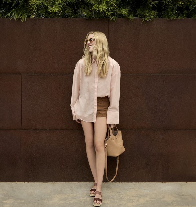 A full shot of a blonde woman in a light pink button-down shirt and brown shorts, posing with a tan purse and sunglasses in an outdoor setting.