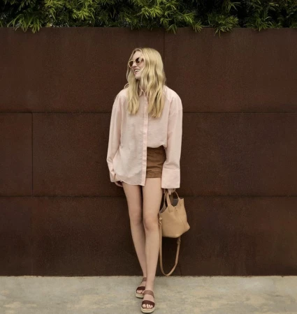A full shot of a blonde woman in a light pink button-down shirt and brown shorts, posing with a tan purse and sunglasses in an outdoor setting.