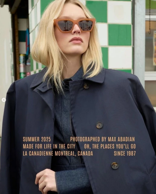 A blonde woman wearing tan sunglasses and a navy blue trench coat poses in front of a green and white tiled background in a medium shot.