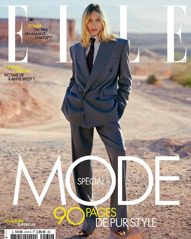 Model Anja Rubik wearing a grey oversized pantsuit and striped shirt standing outdoors in a desert landscape for Elle France magazine cover
