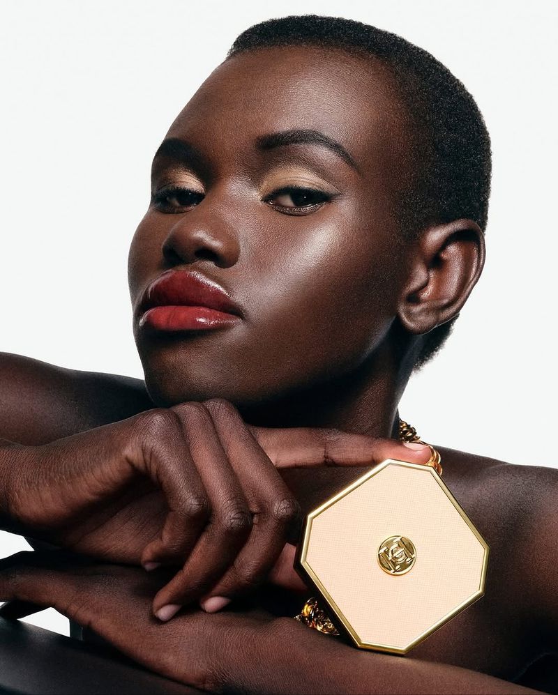 Close-up of a dark-skinned woman with a geometric gold and tan compact, showcasing beauty and fashion in a studio setting.