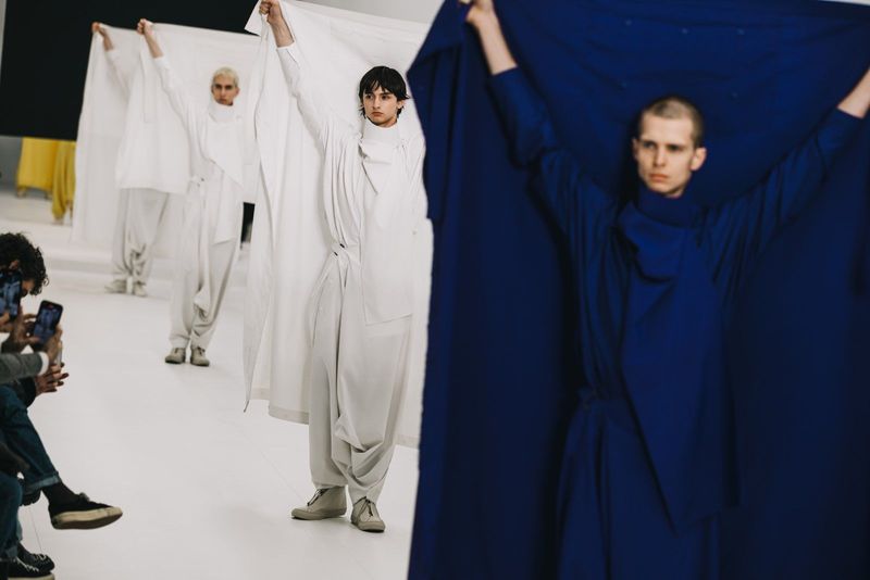 Runway fashion show featuring models in draped white and blue garments, captured in a bright studio with a photographer visible to the side.