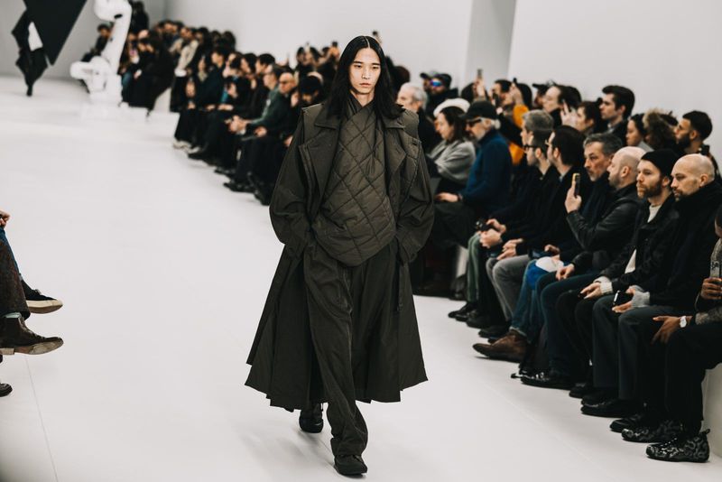 Runway model in a dark green quilted vest, matching long coat, and pants walks toward the camera during a fashion show with audience members.