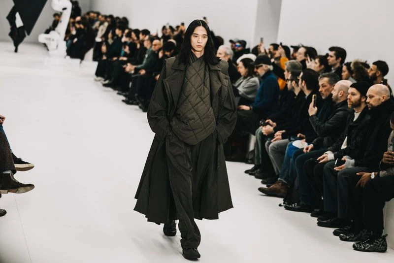 Runway model in a dark green quilted vest, matching long coat, and pants walks toward the camera during a fashion show with audience members.