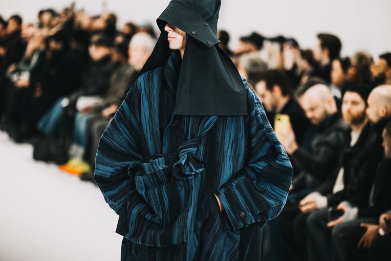 Runway fashion show featuring a model in a striped coat and black head covering, with a crowd of spectators in the background.