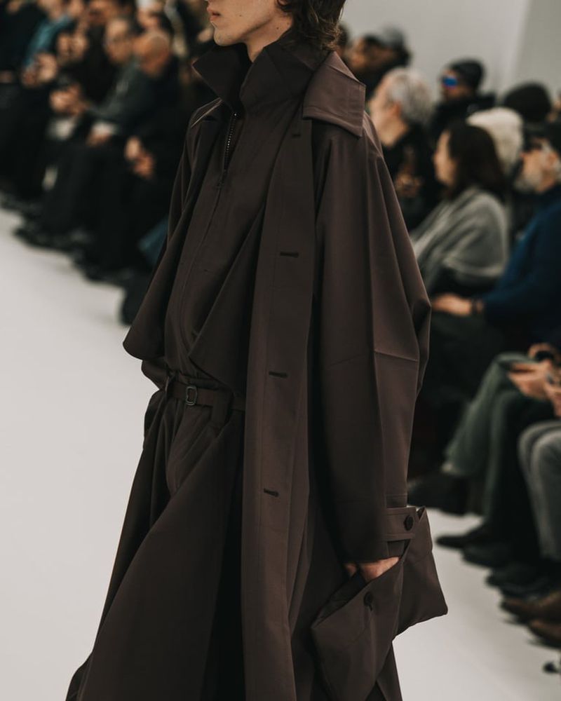 Fashion show runway featuring a model in a brown oversized coat, wide-legged trousers, and a high-collared shirt, surrounded by blurred audience.