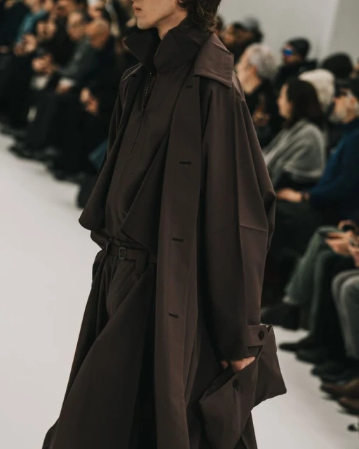 Fashion show runway featuring a model in a brown oversized coat, wide-legged trousers, and a high-collared shirt, surrounded by blurred audience.