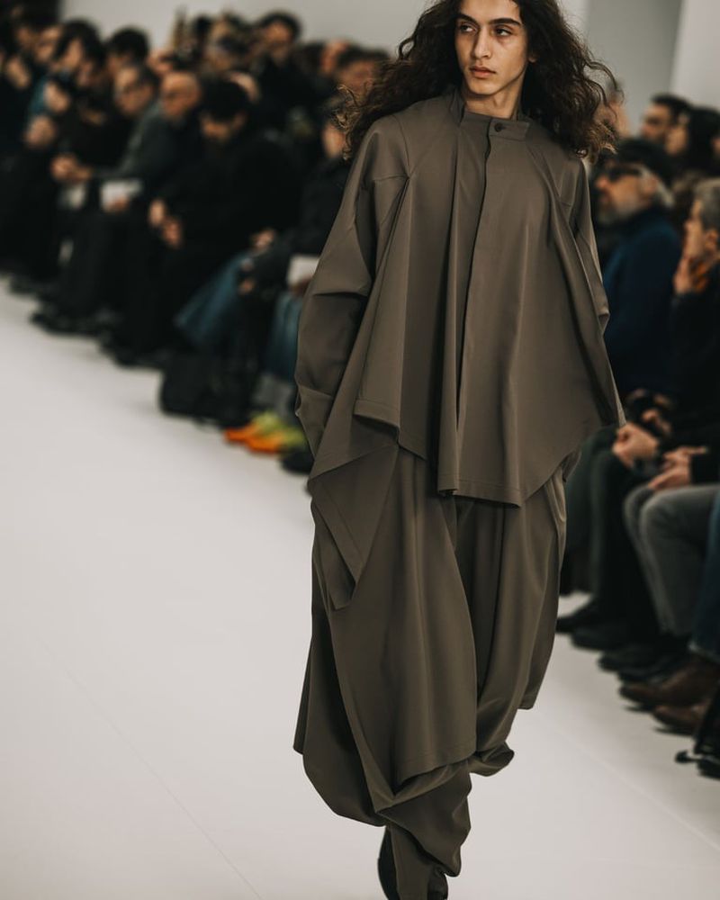 Runway fashion show featuring a model in a loose-fitting grey two-piece outfit walking on a white runway, audience members in the background.