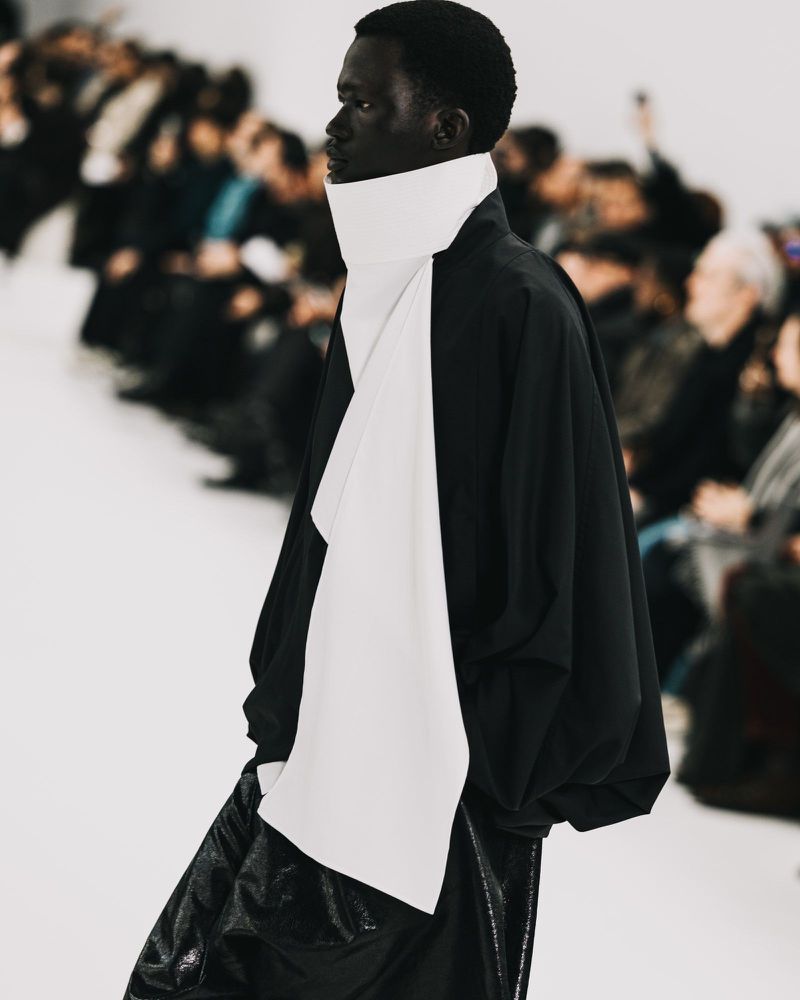Runway fashion show featuring a person of color wearing a black top and shiny black skirt; audience visible in the background.
