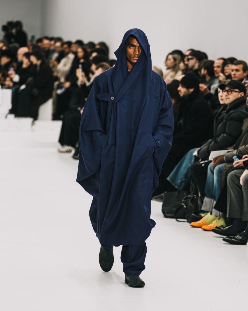 A fashion model on a runway at a show wearing a dark blue hooded robe-like coat and loose pants as an audience watches in the background.