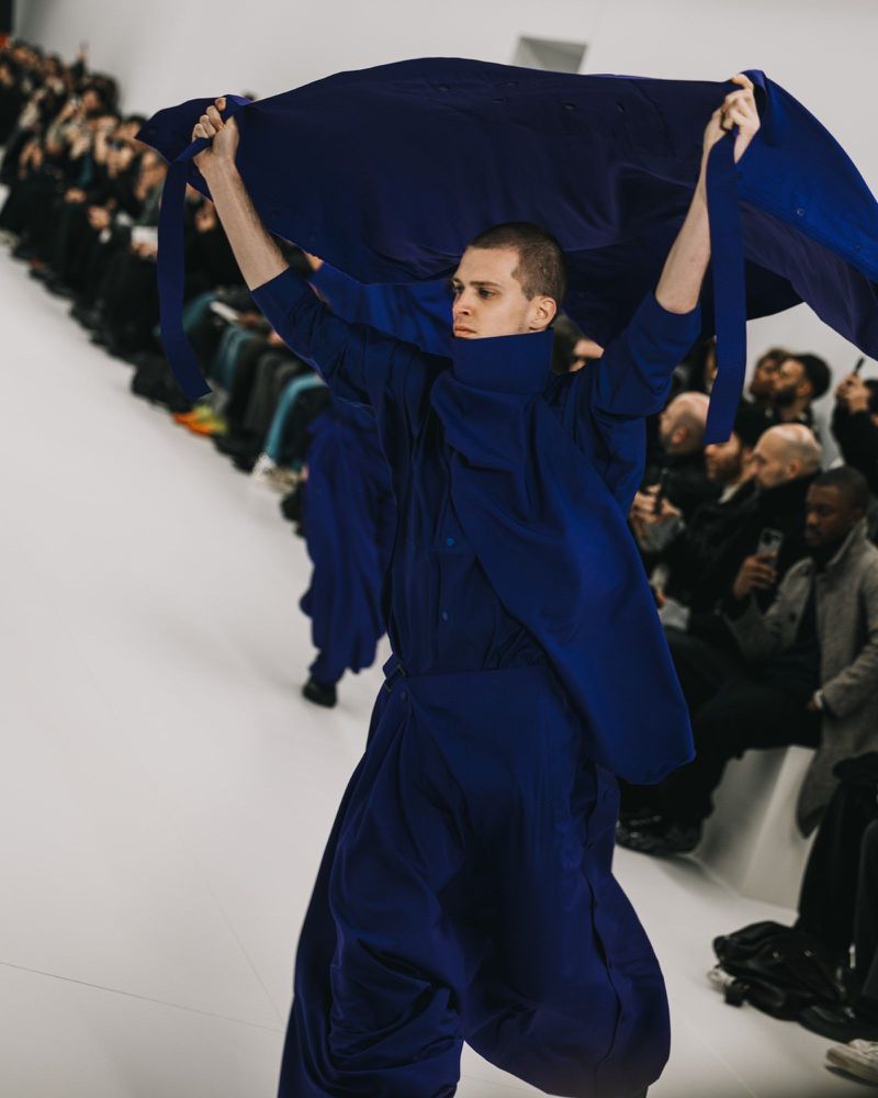 Runway show during fashion week: A male fashion model carries a navy blue fabric on a white runway while other models walk behind him.