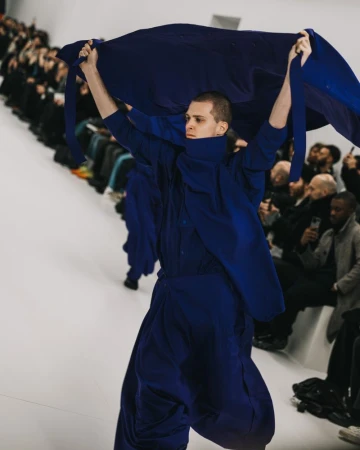 Runway show during fashion week: A male fashion model carries a navy blue fabric on a white runway while other models walk behind him.