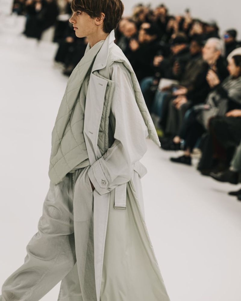Runway fashion show featuring an adult model walking in a light grey oversized trench coat, quilted jacket, and pants, with a crowd watching.