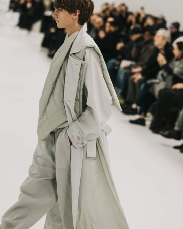 Runway fashion show featuring an adult model walking in a light grey oversized trench coat, quilted jacket, and pants, with a crowd watching.