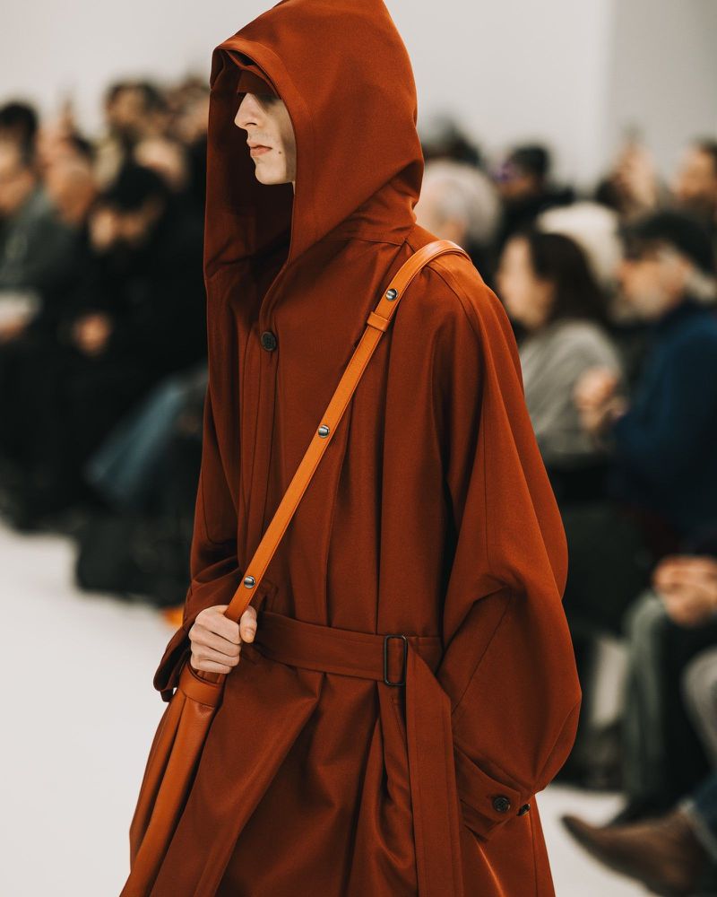 Runway fashion show: A model walks in a hooded coat with a belted waist and shoulder bag at a fashion show with a blurred audience.