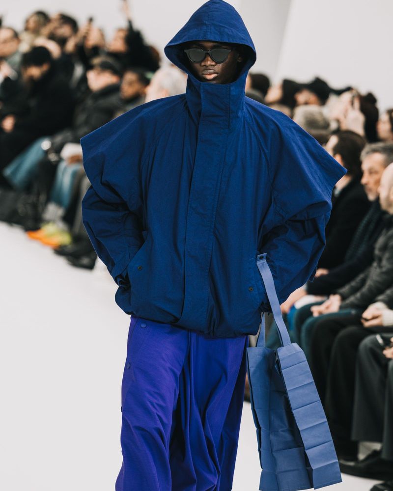 Model wearing a blue hooded jacket and matching trousers carrying a blue bag on a runway with seated audience members