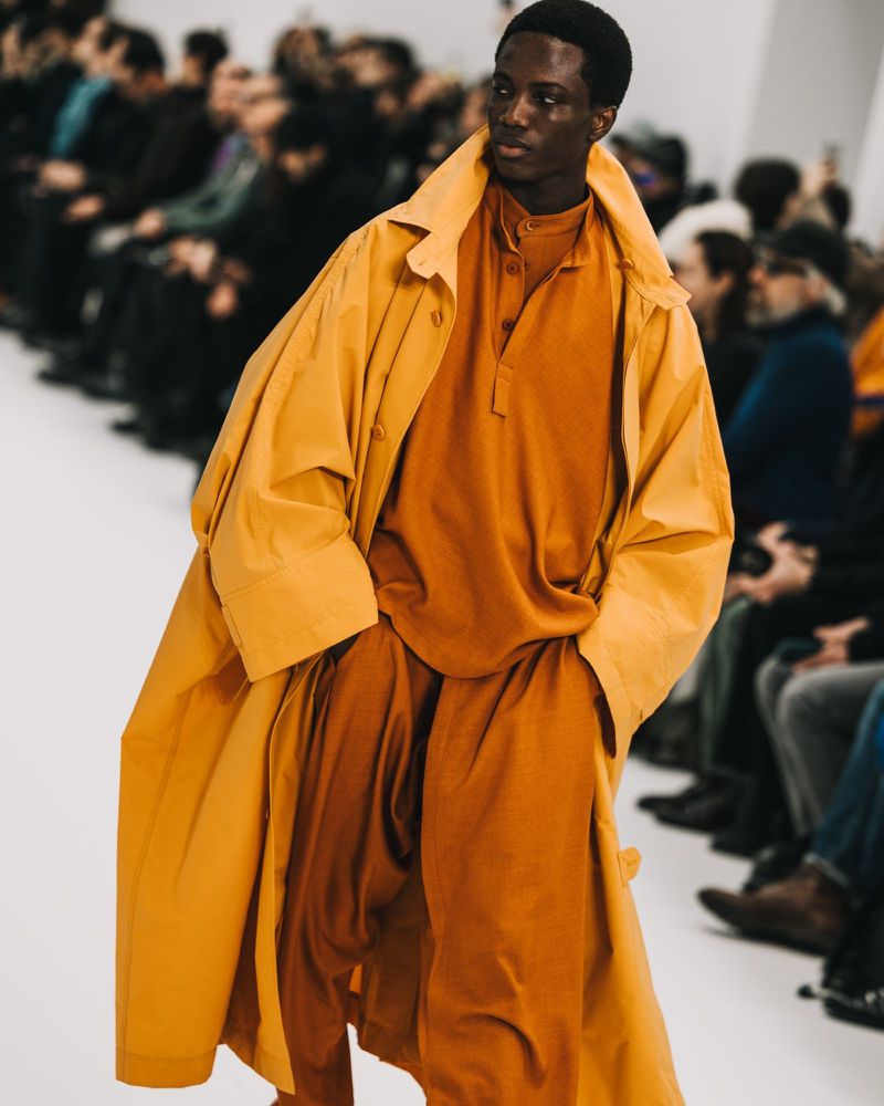 Runway fashion show featuring a model wearing a mustard coat and matching outfit on a white catwalk, audience seated in background.