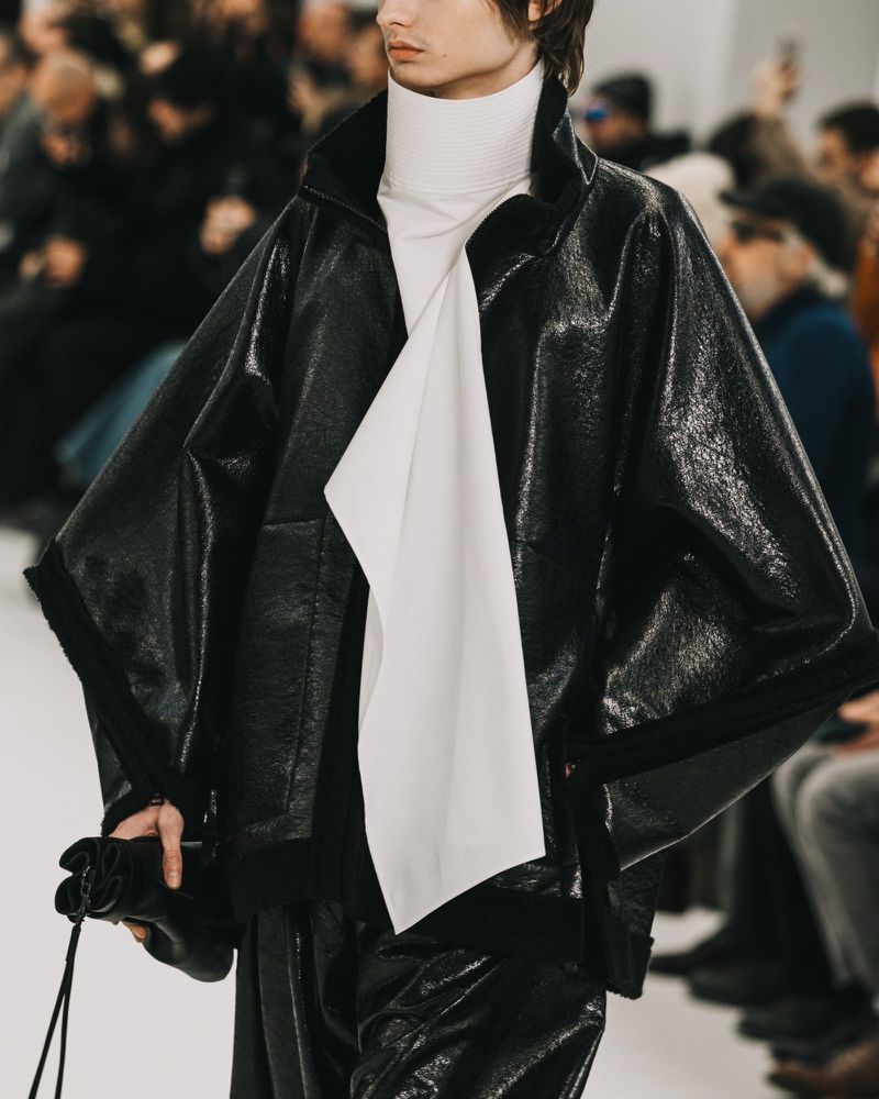 Runway fashion presentation featuring an adult male model in a black leather jacket and pants with a white turtle neck top.