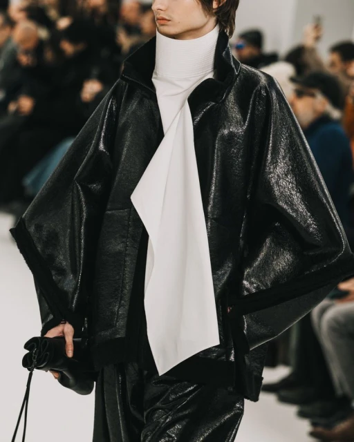 Runway fashion presentation featuring an adult male model in a black leather jacket and pants with a white turtle neck top.