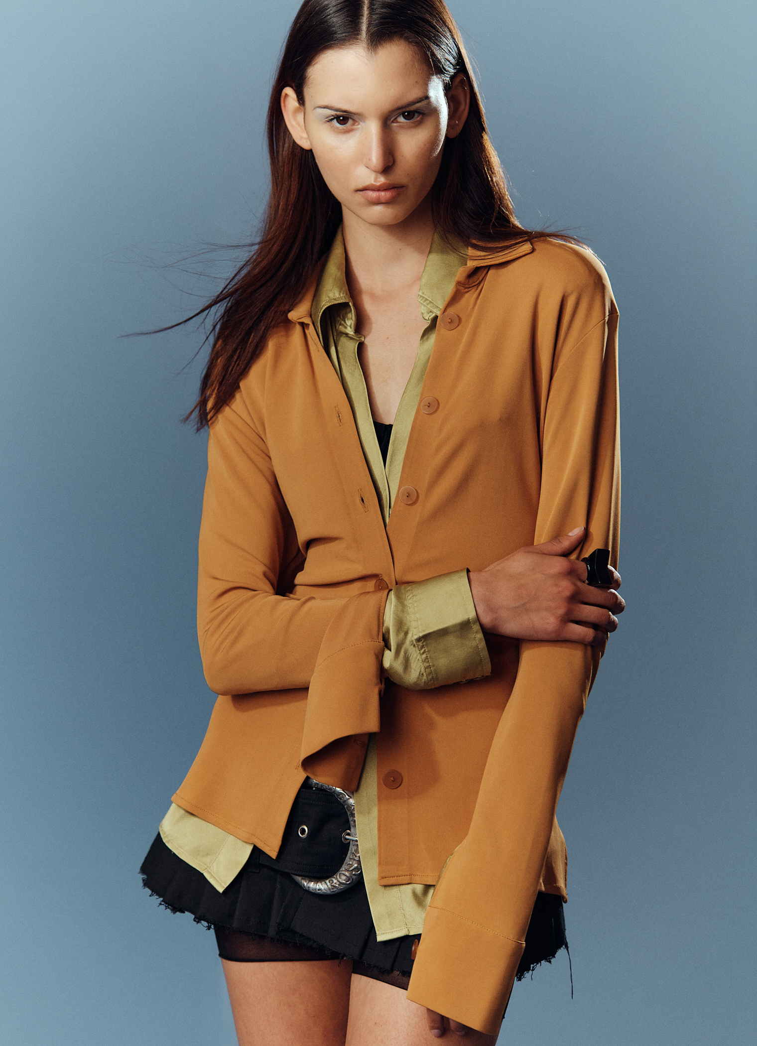 Model Larissa Schumacher in a mustard-brown button-down shirt and layered olive blouse against a plain grey studio backdrop