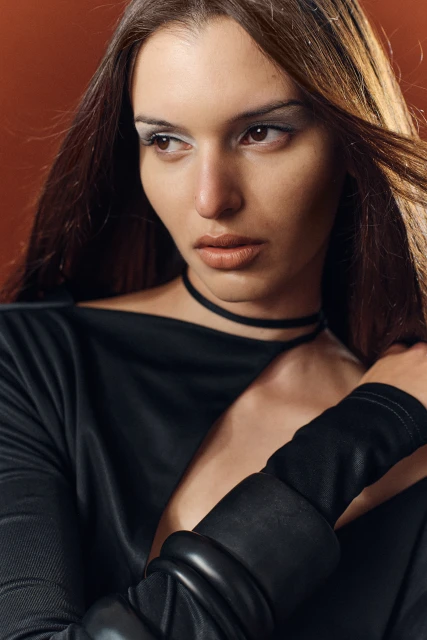 Portrait of a young woman with dramatic makeup and dark clothing in front of a brick-red backdrop. The woman's gaze is averted.