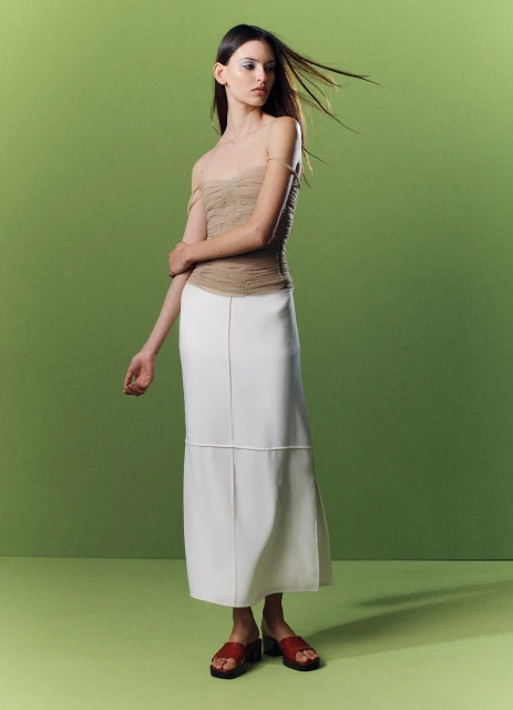 A woman models a beige top and white midi skirt against a gradient green background in a fashion studio shot with a full figure view.