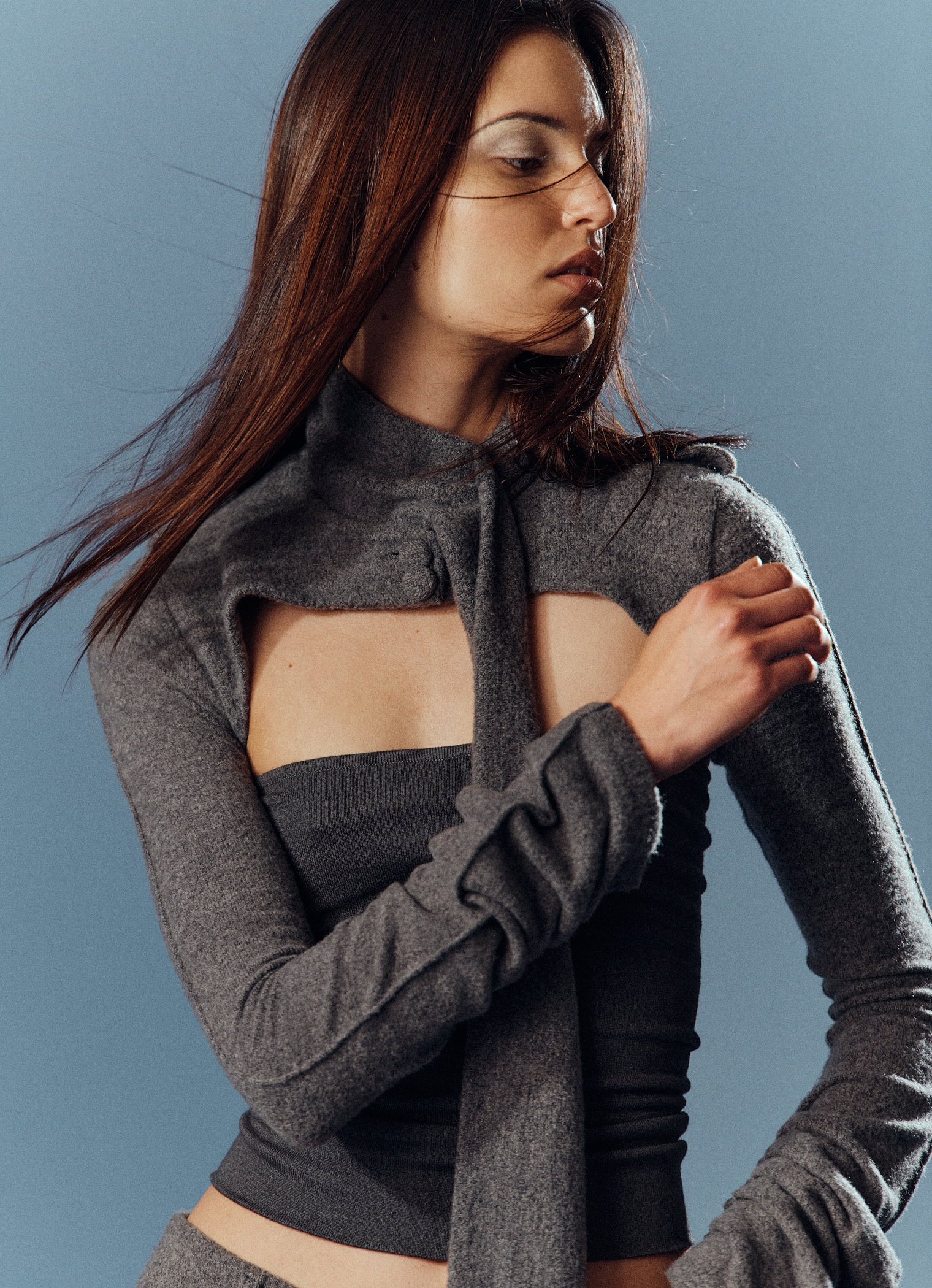 A fashionable woman in a grey knit top and pants poses against a blue backdrop, showcasing a minimalist style and elegant design.