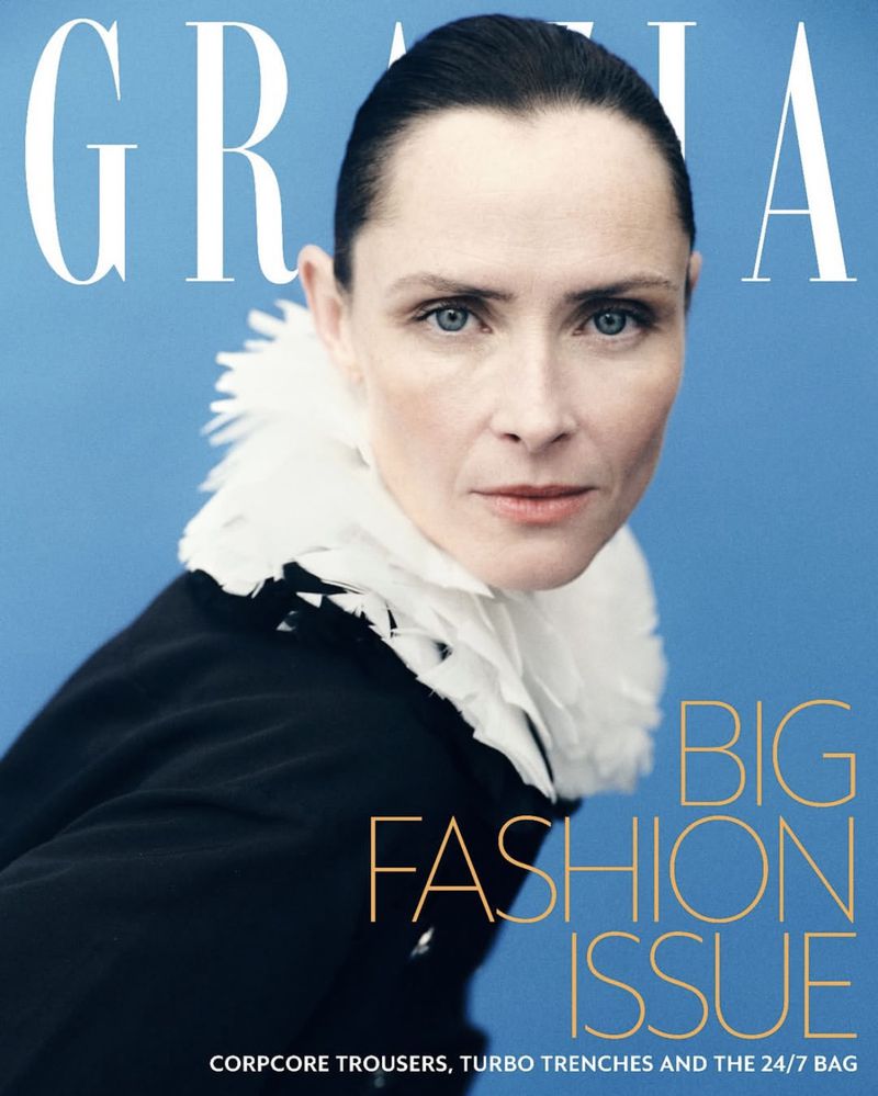 Fashion magazine cover featuring a woman with slicked-back hair, wearing a dark coat with a white collar on a light blue background.