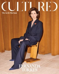 Fernanda Torres seated on a wooden stool wearing a dark pinstripe pantsuit against a gold curtain backdrop