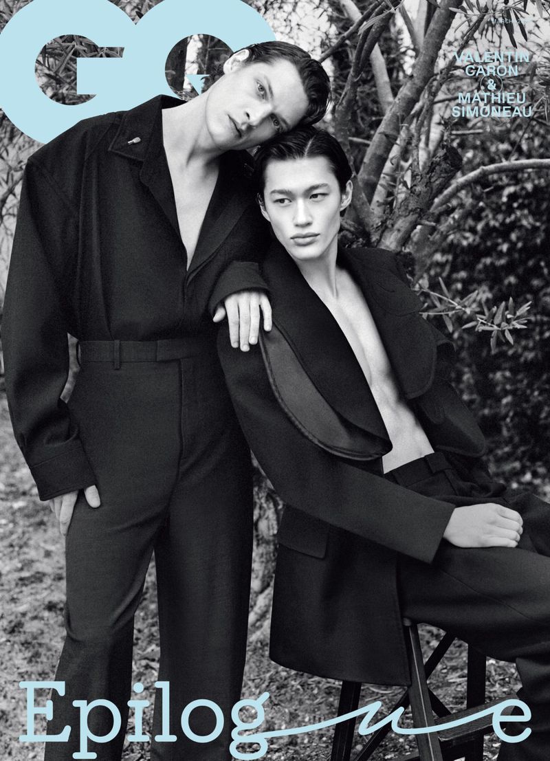 Two adult males pose in black jackets outdoors for a GQ magazine cover, standing by a tree in a black and white fashion editorial.