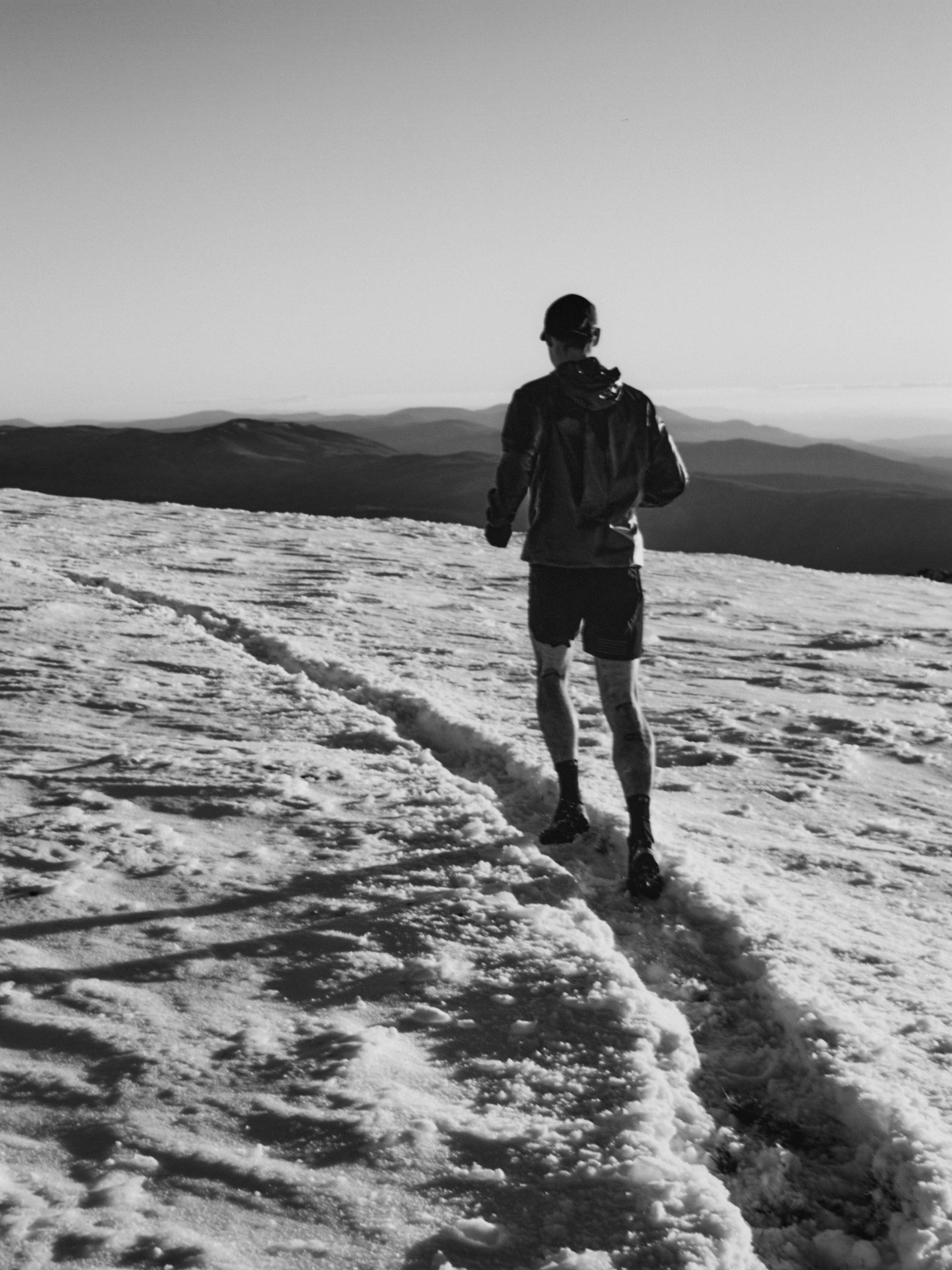 A man runs on a snowy mountain path under a clear sky, captured from a low angle, showing a serene, black and white outdoor scene.