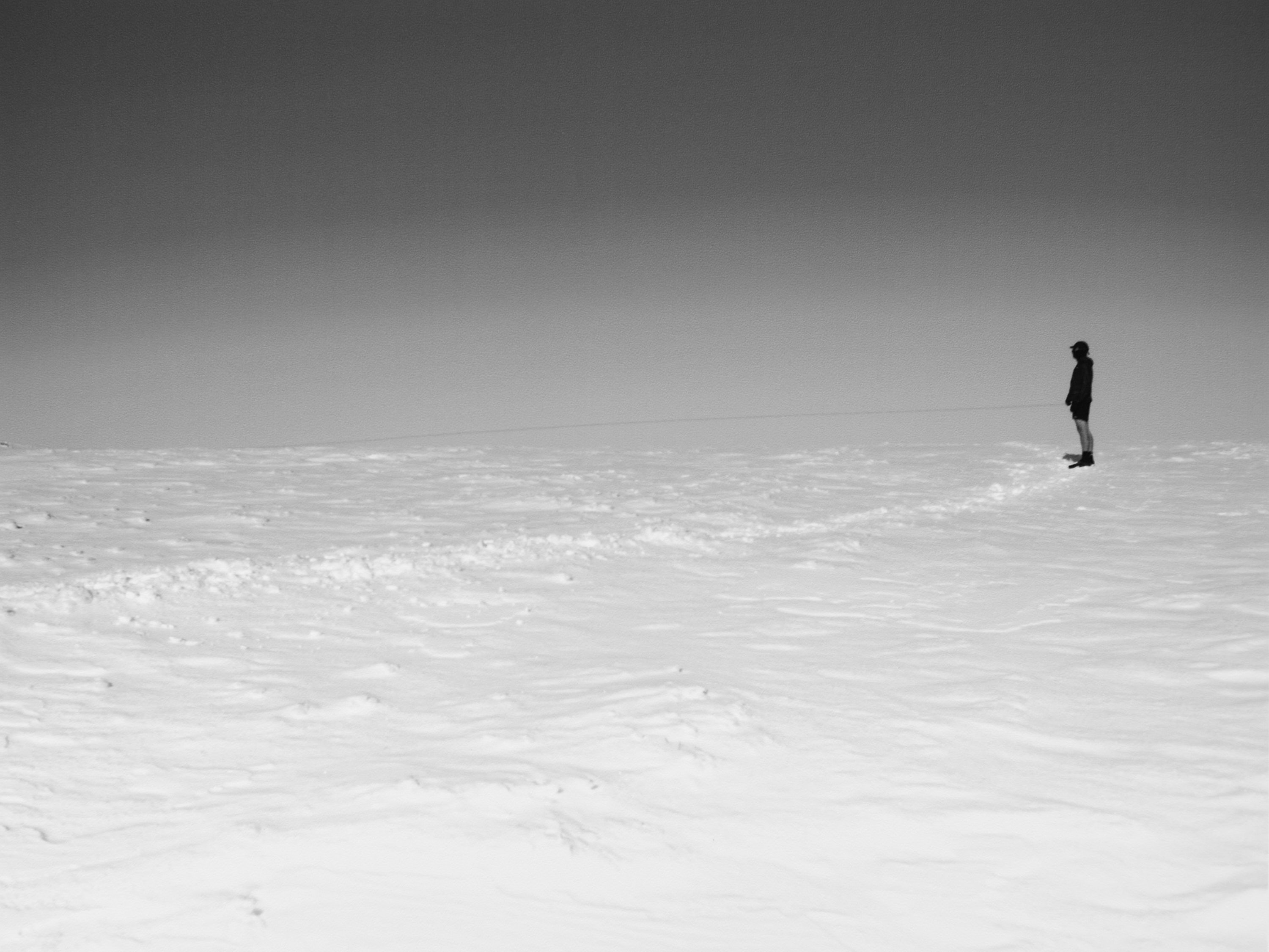 Monochrome landscape featuring a distant, solitary figure standing on a snow-covered field under a grey sky, emphasizing isolation.