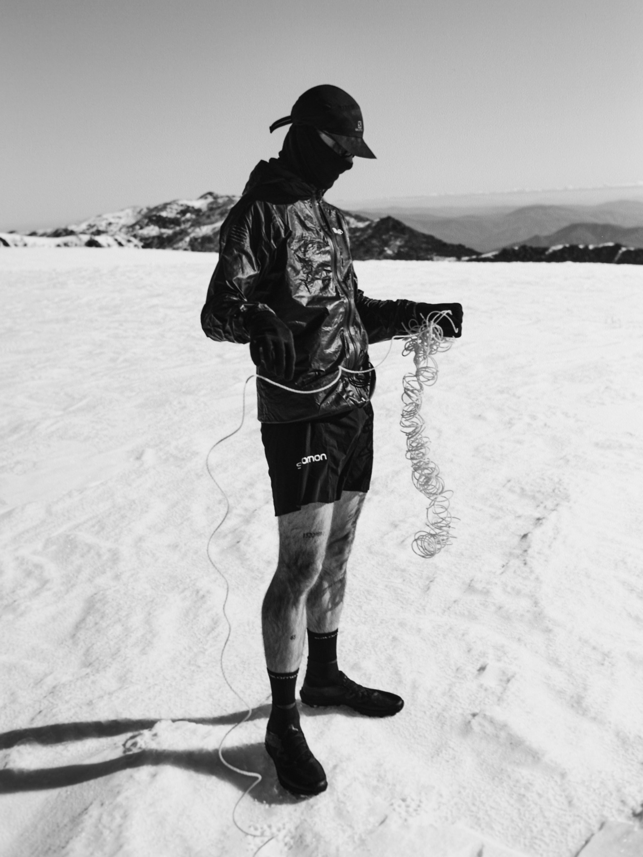 Black and white shot of an adult wearing athletic wear outdoors in a snowy environment, coiling a white rope on a bright, sunny day.