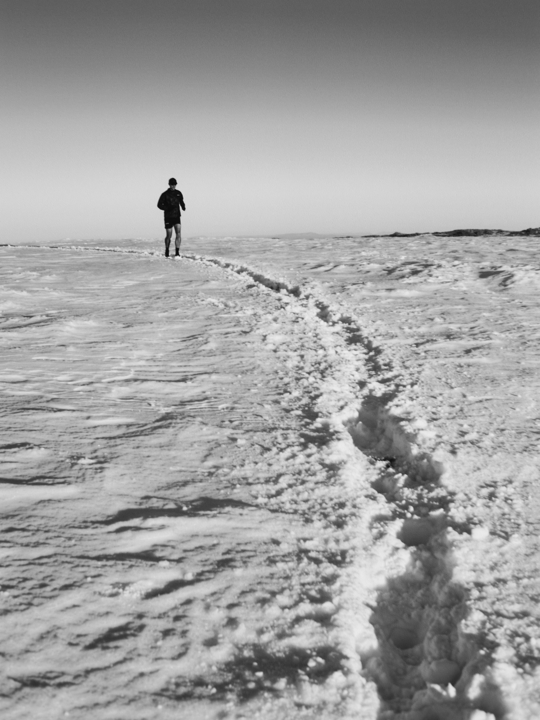 Monochrome image of a man walking alone on a snowy landscape, leaving a trail of footprints, under a clear sky, creating a stark, remote scene.