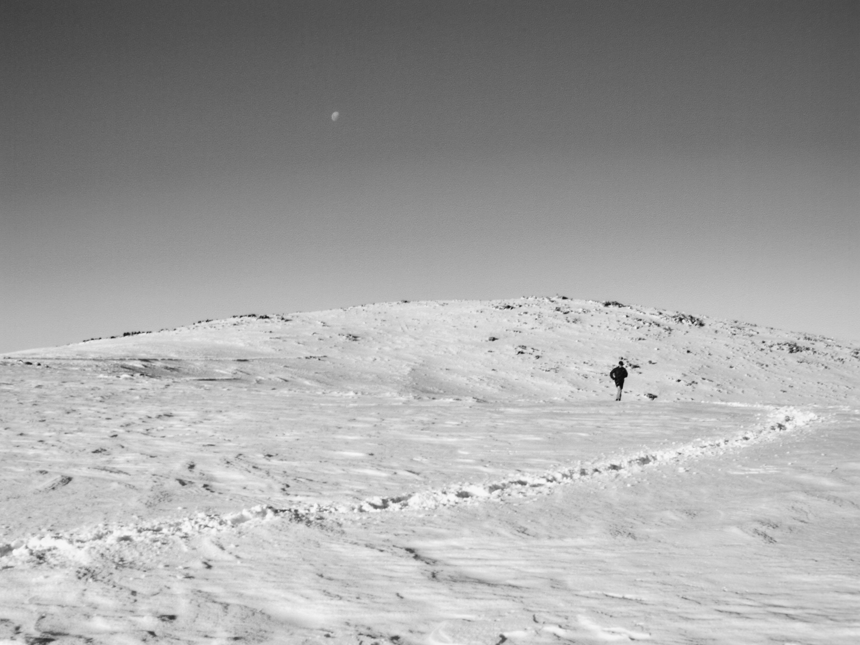 Monochrome landscape features a lone figure walking across a snowy field toward a hill under a bright sky with a crescent moon.