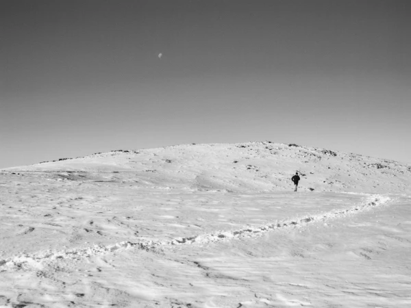 Monochrome landscape features a lone figure walking across a snowy field toward a hill under a bright sky with a crescent moon.