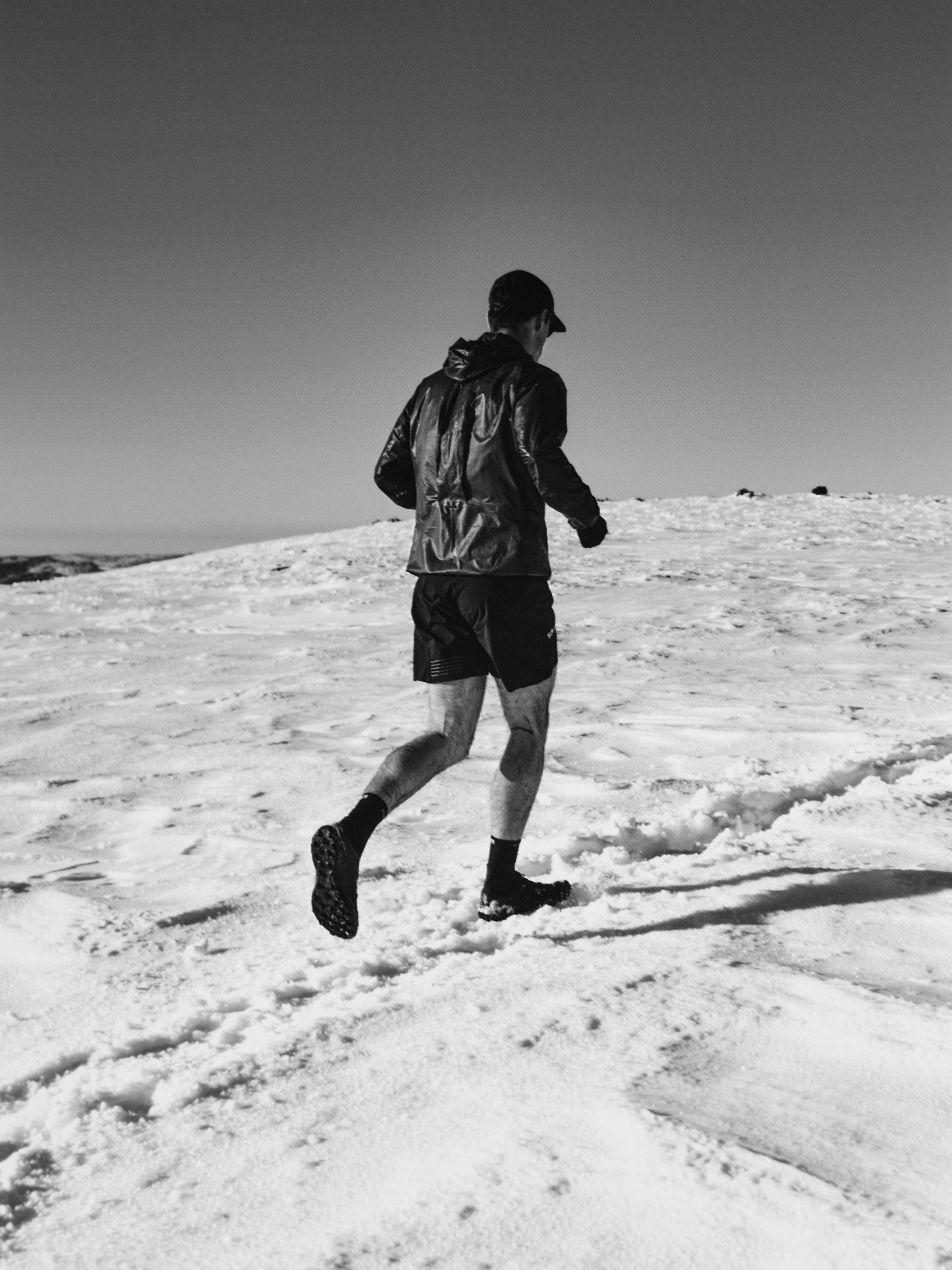 Monochrome shot of a person running across a snow-covered landscape, wearing athletic wear and a cap, under a clear sky. 