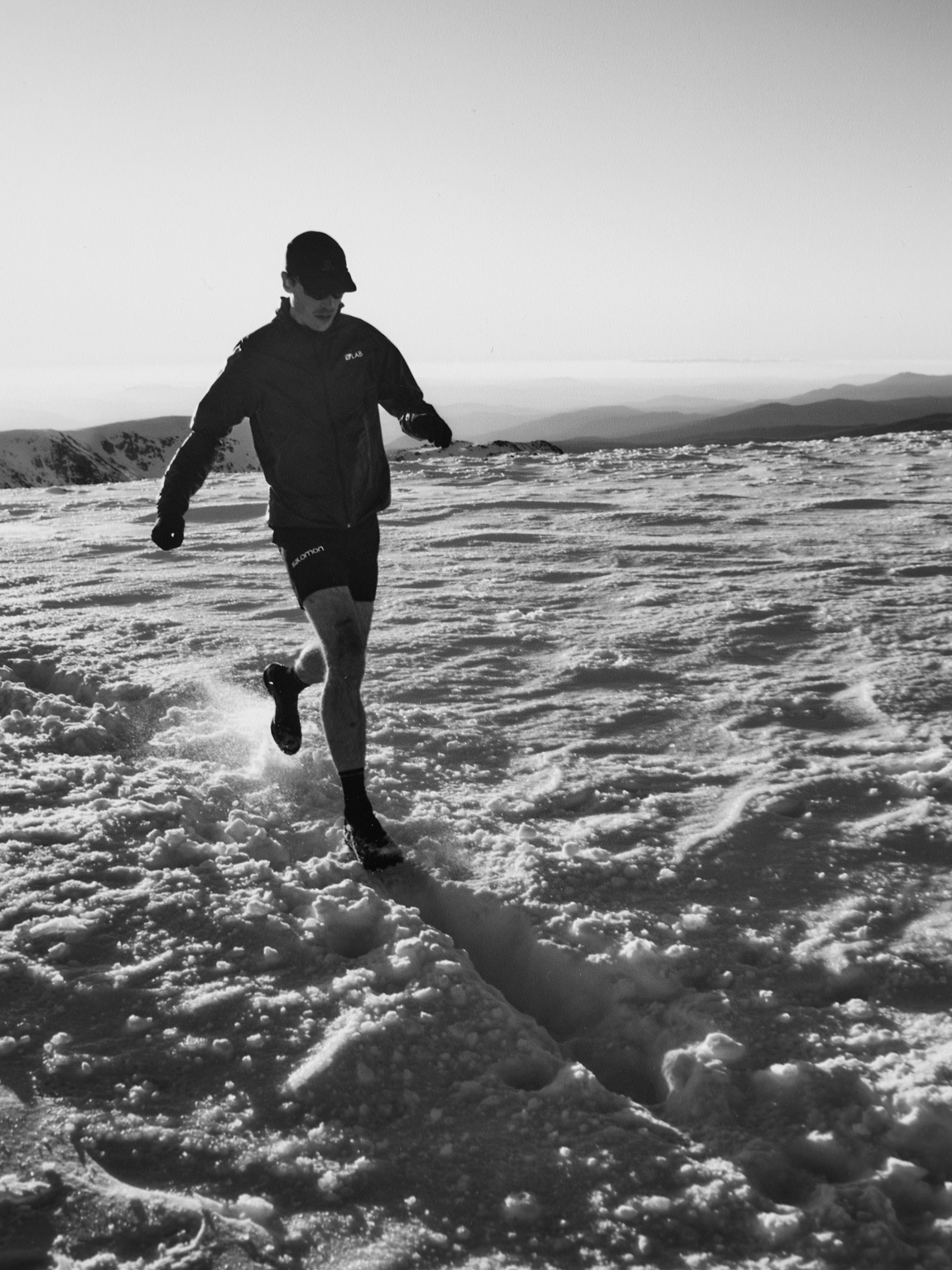 Monochrome shot of a male athlete running on snow-covered mountain, wearing athletic attire and a cap, with a backdrop of distant peaks.