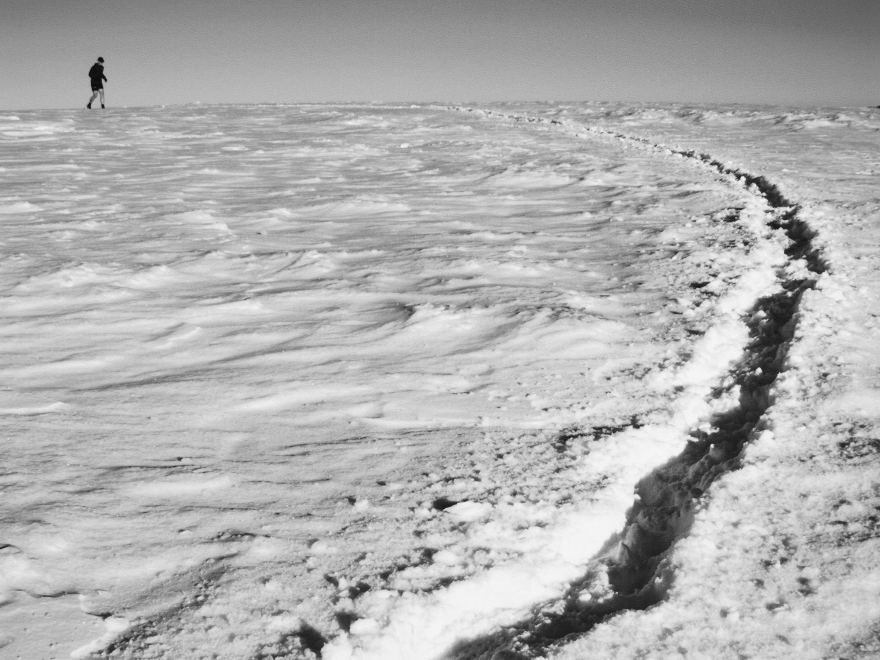 Person walking on vast snow-covered field, with a winding track of footprints leading into the distance under a clear sky.