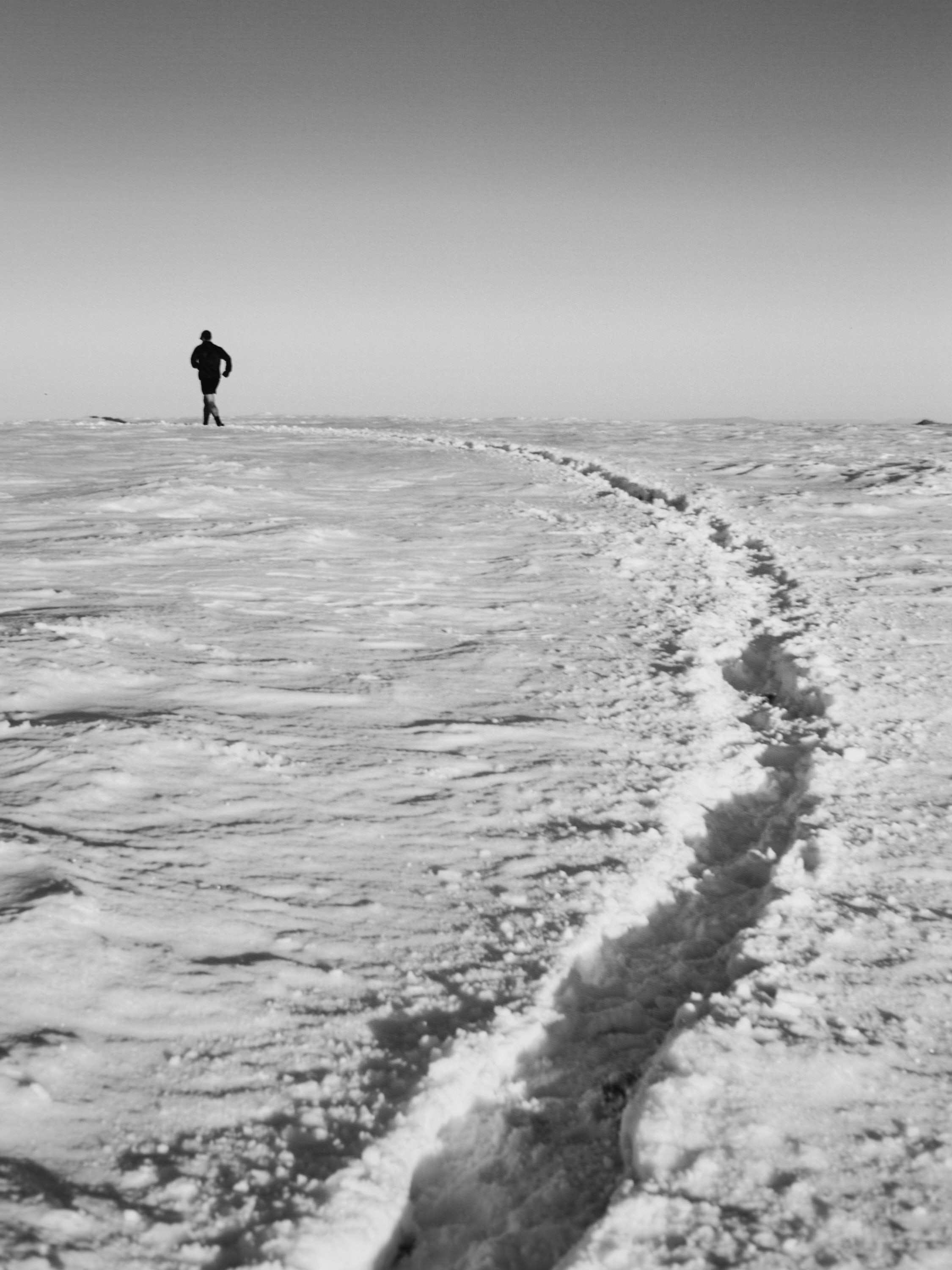 A solitary figure runs across a snow-covered landscape, leaving a trail of footprints behind him, under a light sky.