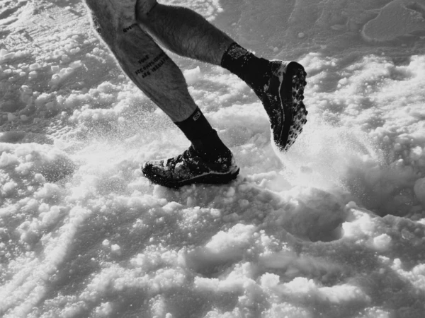Monochrome shot of a person's legs running in snow, wearing athletic shoes and black socks with tattoo on the calf.