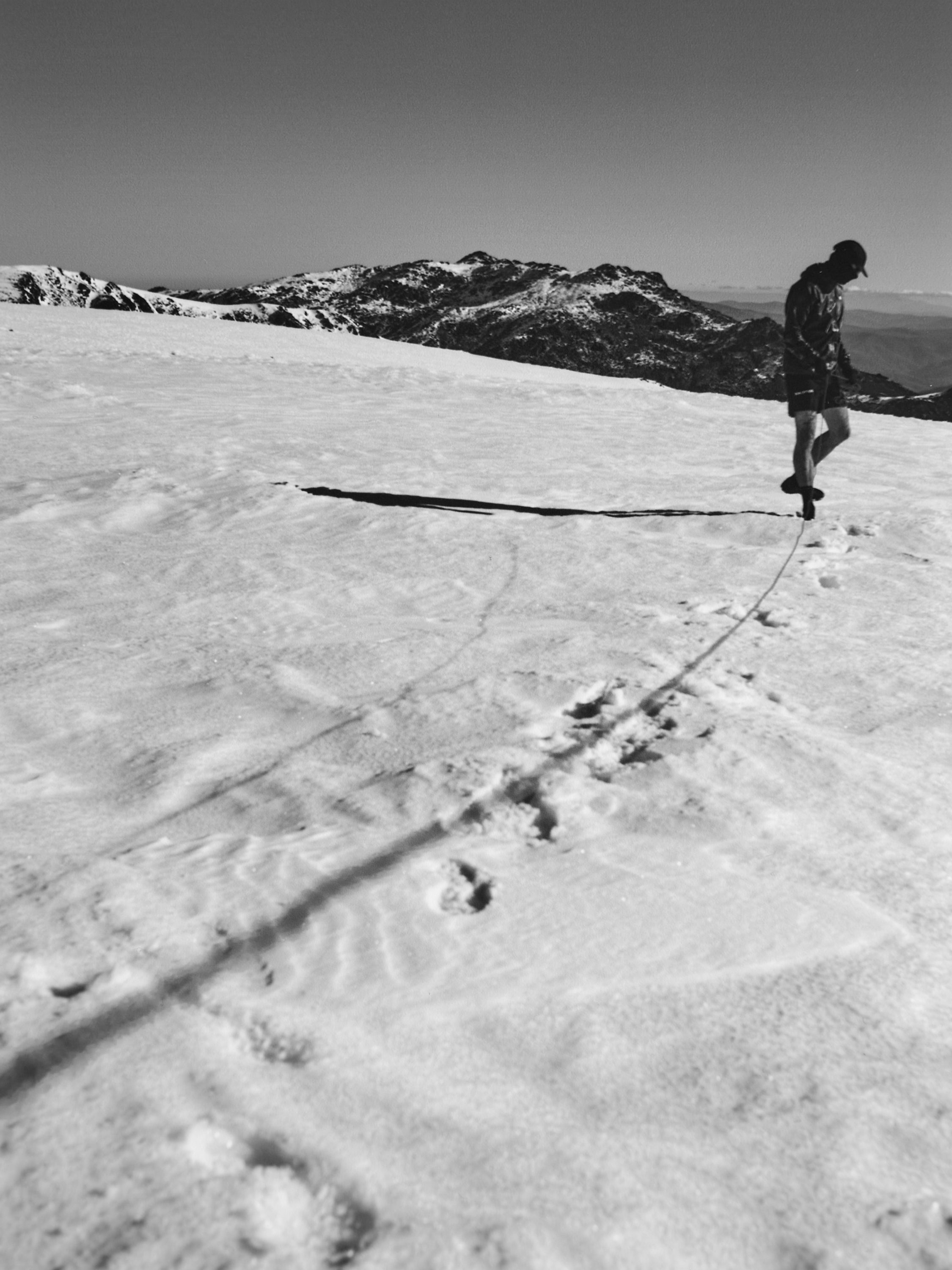Monochrome shot of a person walking across a snow-covered landscape with mountains in the background, casting a long shadow.