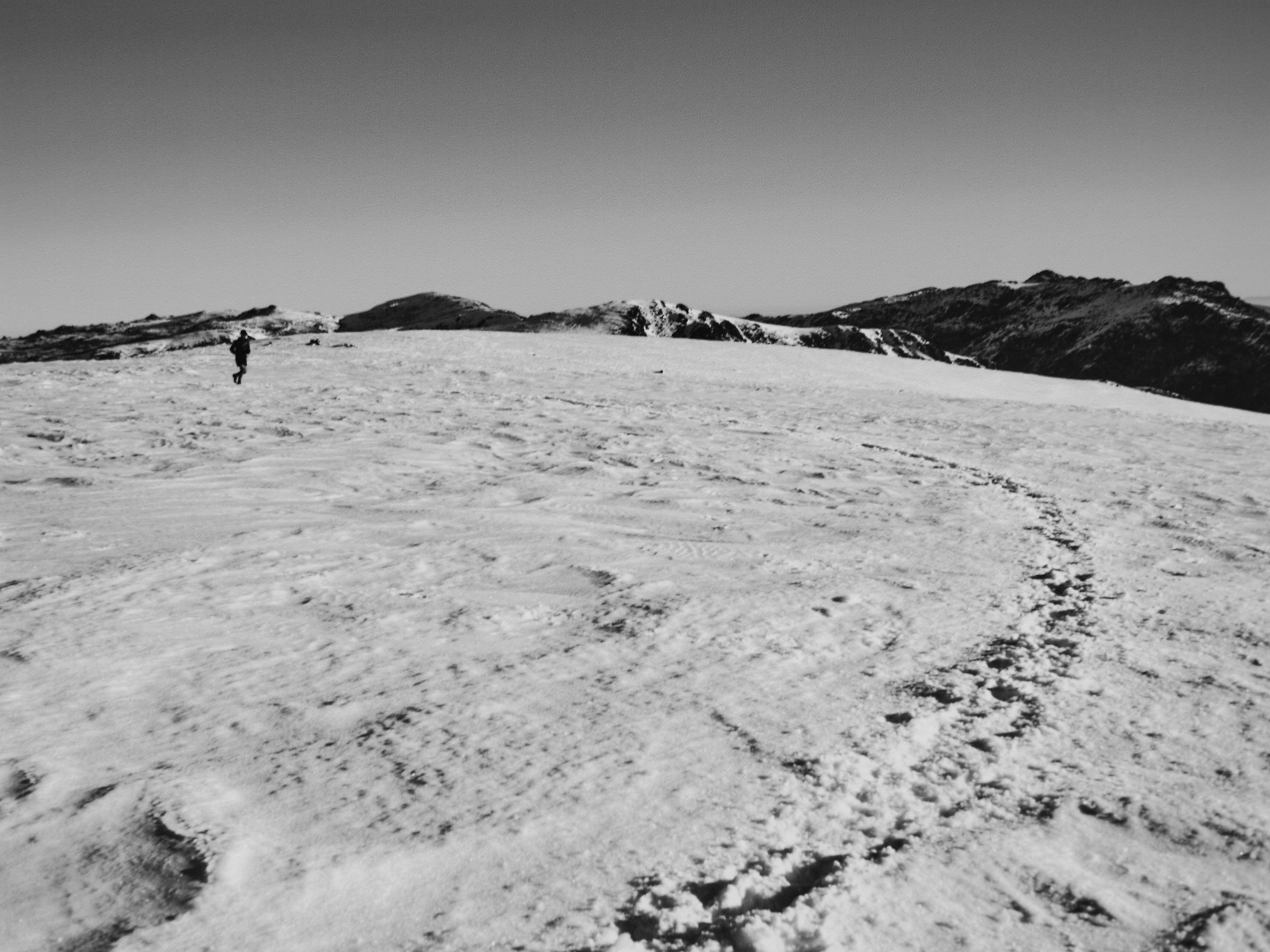 Monochrome image of a lone adult walking across a snow-covered mountain landscape under a clear sky with visible footprints.